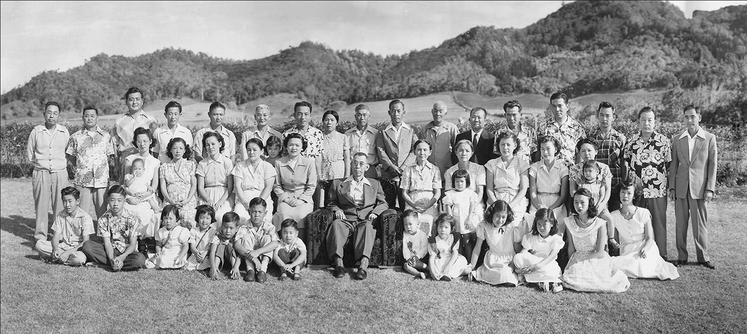 Multi-generation family portrait outdoors on a grassy lawn, with grandparents seated in the center and children and adults gathered around against a backdrop of hills and trees.