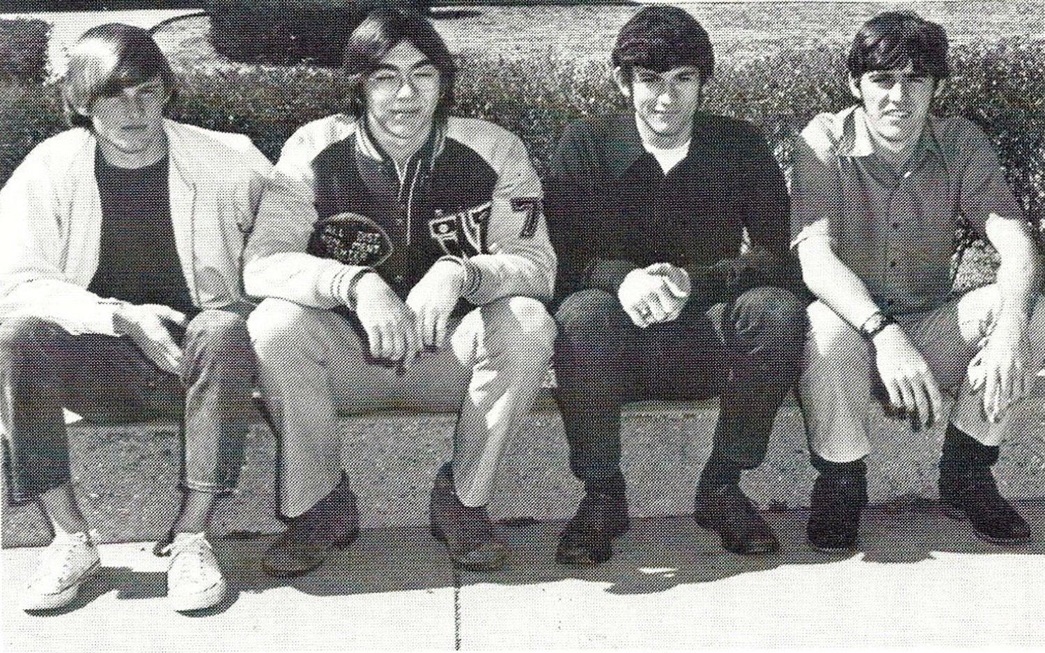Five young men sit closely on a low wall outdoors, facing the camera in casual jackets and jeans.