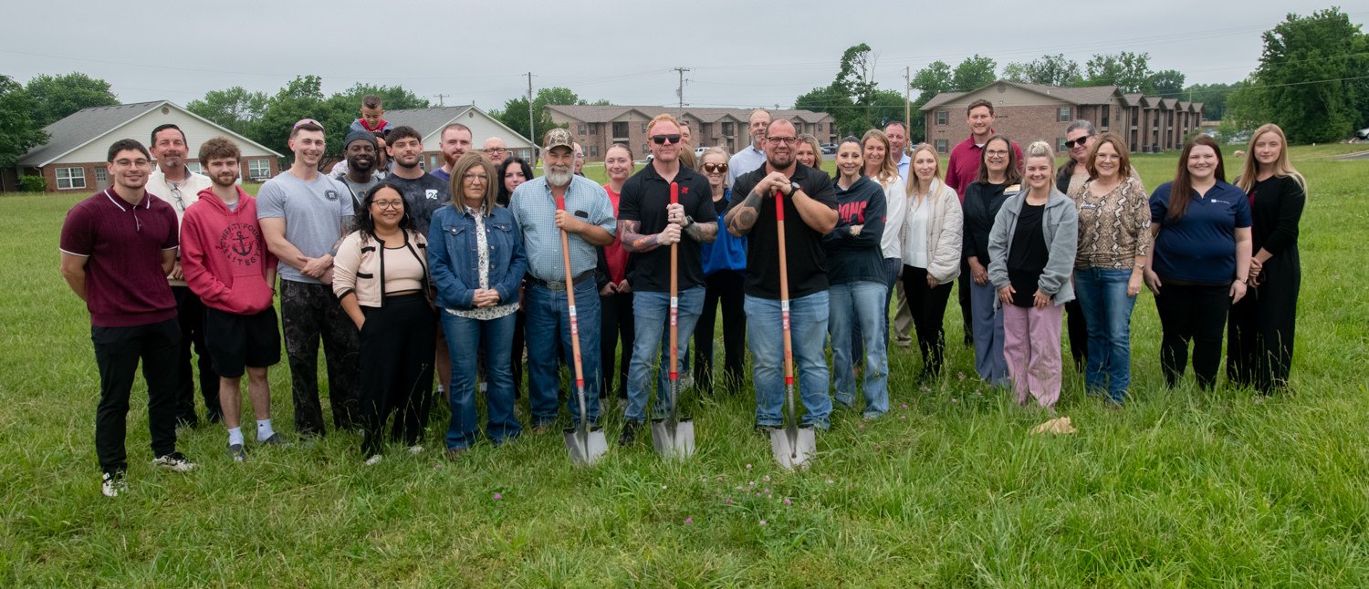 Group of people posing with shovels in a grassy field, likely members at a ground-breaking or community project event with houses in the background.