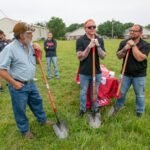 Group of men with shovels at a ground-breaking event in a grassy field, preparing to dig while others watch nearby.