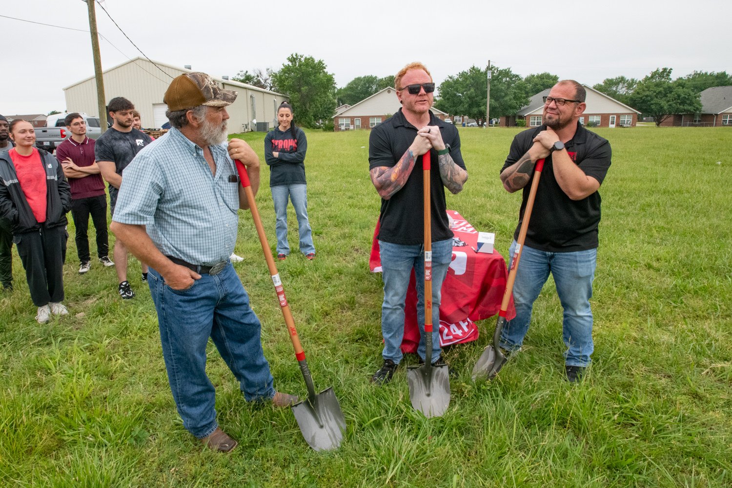 Group of men with shovels at a ground-breaking event in a grassy field, preparing to dig while others watch nearby.