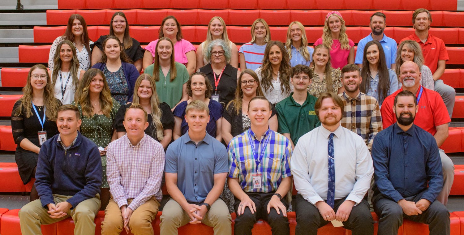 Large group of students and staff posing for a portrait on red gym bleachers indoors/at a school or organization event, smiling at the camera.
