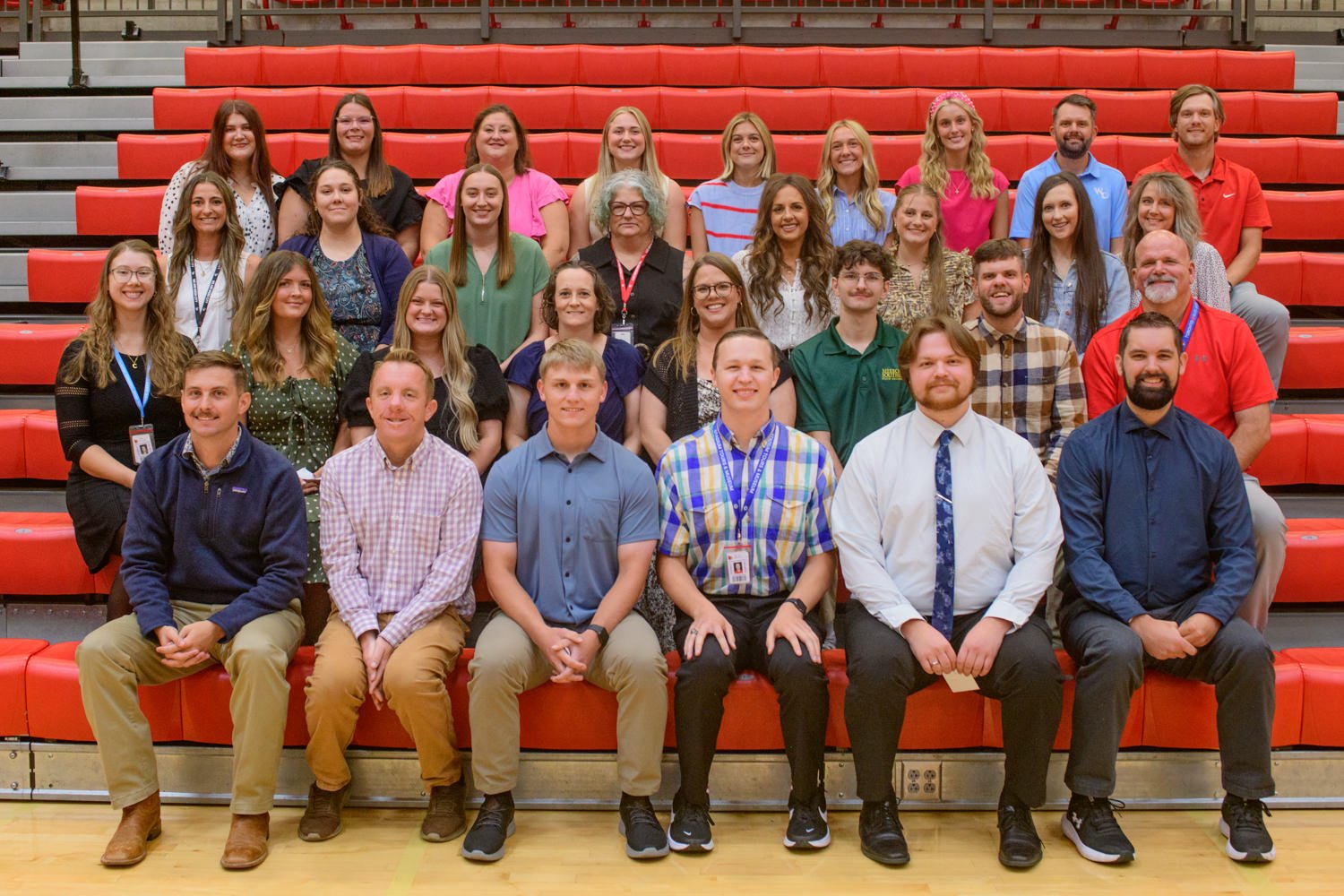 Large group of students and staff posed on red bleachers in a gymnasium. Front row seated, back rows standing.