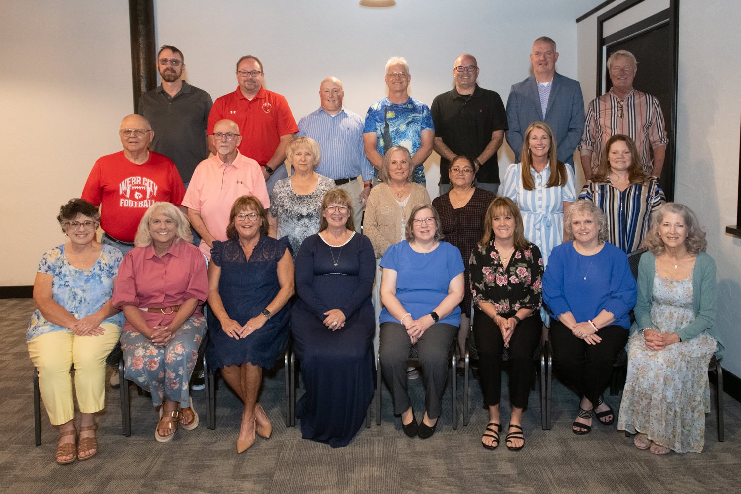 Three-row group portrait of about 20 adults in a meeting room, smiling and posing for the photo in casual to business-casual attire.