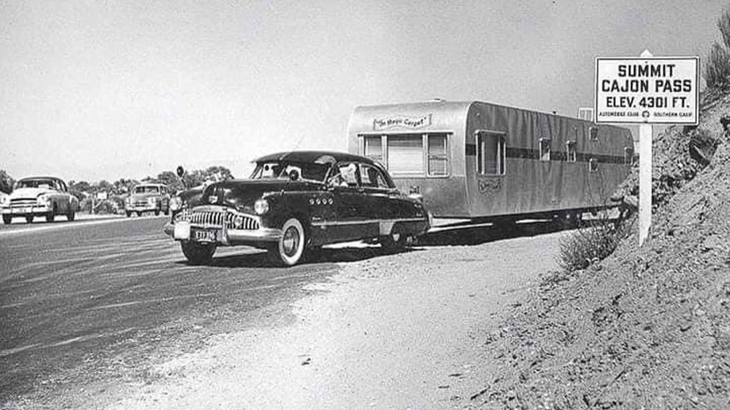 Vintage car towing a travel trailer on a mountain highway, Cajon Pass summit sign visible in the background.