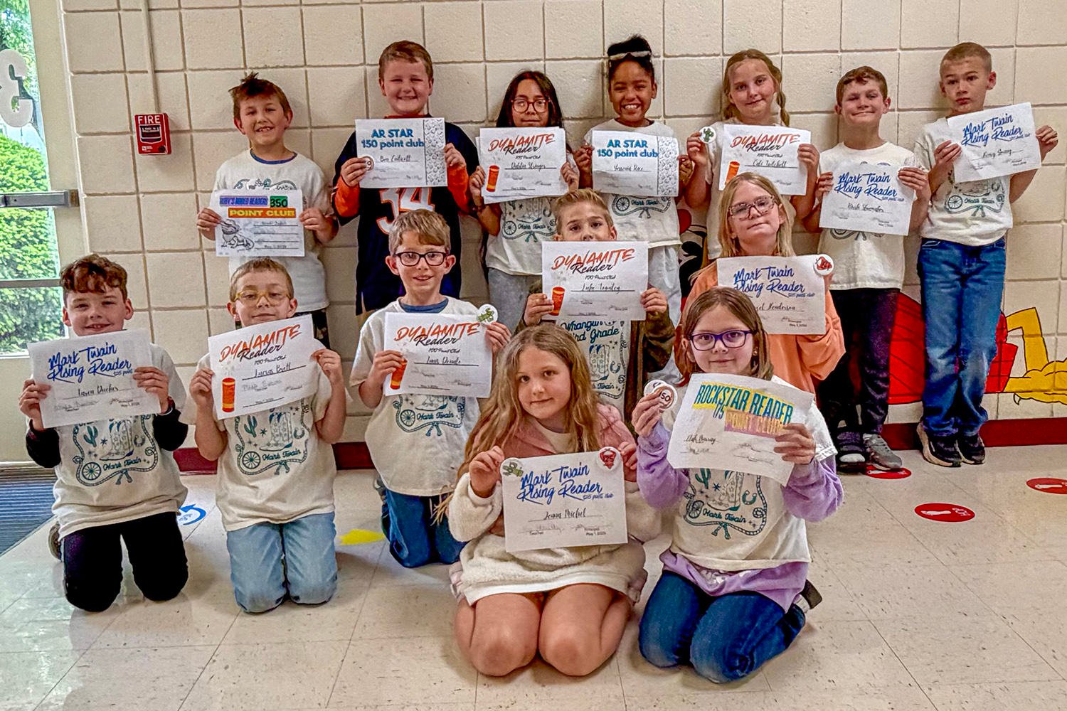 Group of elementary school children in a hallway, kneeling and standing, proudly holding certificates for a reading program and smiling at the camera.