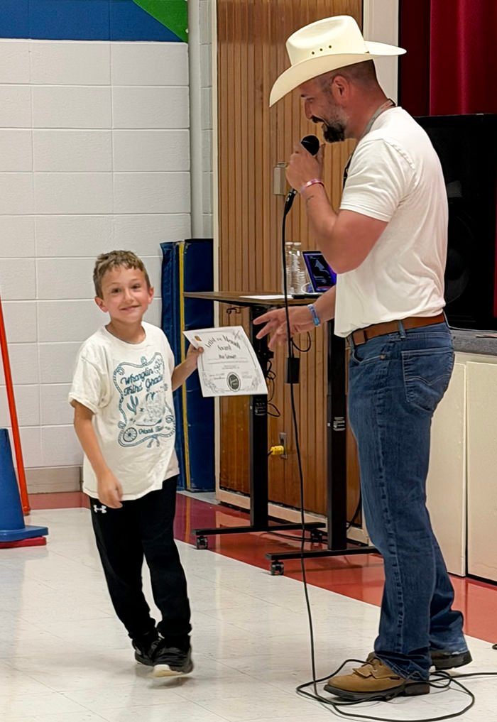 Man in a cowboy hat hands a certificate to a smiling boy during a school assembly or event in a gym.