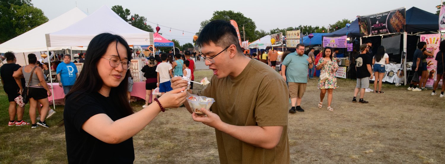 Two friends at an outdoor festival share a dessert, one feeding the other from a bowl as tents and crowds loom in the background.
