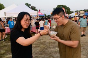 Woman hands a dessert cup to a smiling man at an outdoor festival with tents and crowds in the background.