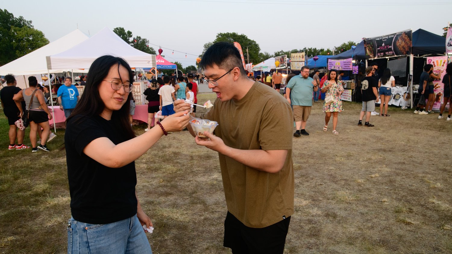 A man in a brown shirt feeds a spoonful of dessert to a woman in a black shirt at a crowded outdoor festival with vendor tents in the background.
