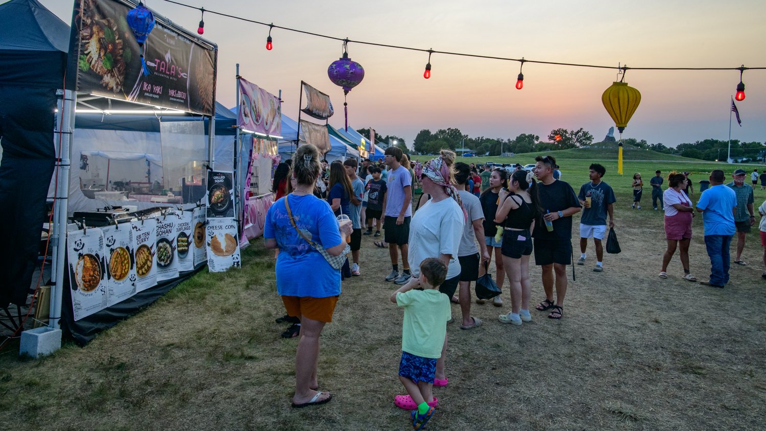 Outdoor festival at dusk with food stalls and string lights, people lining up for meals.