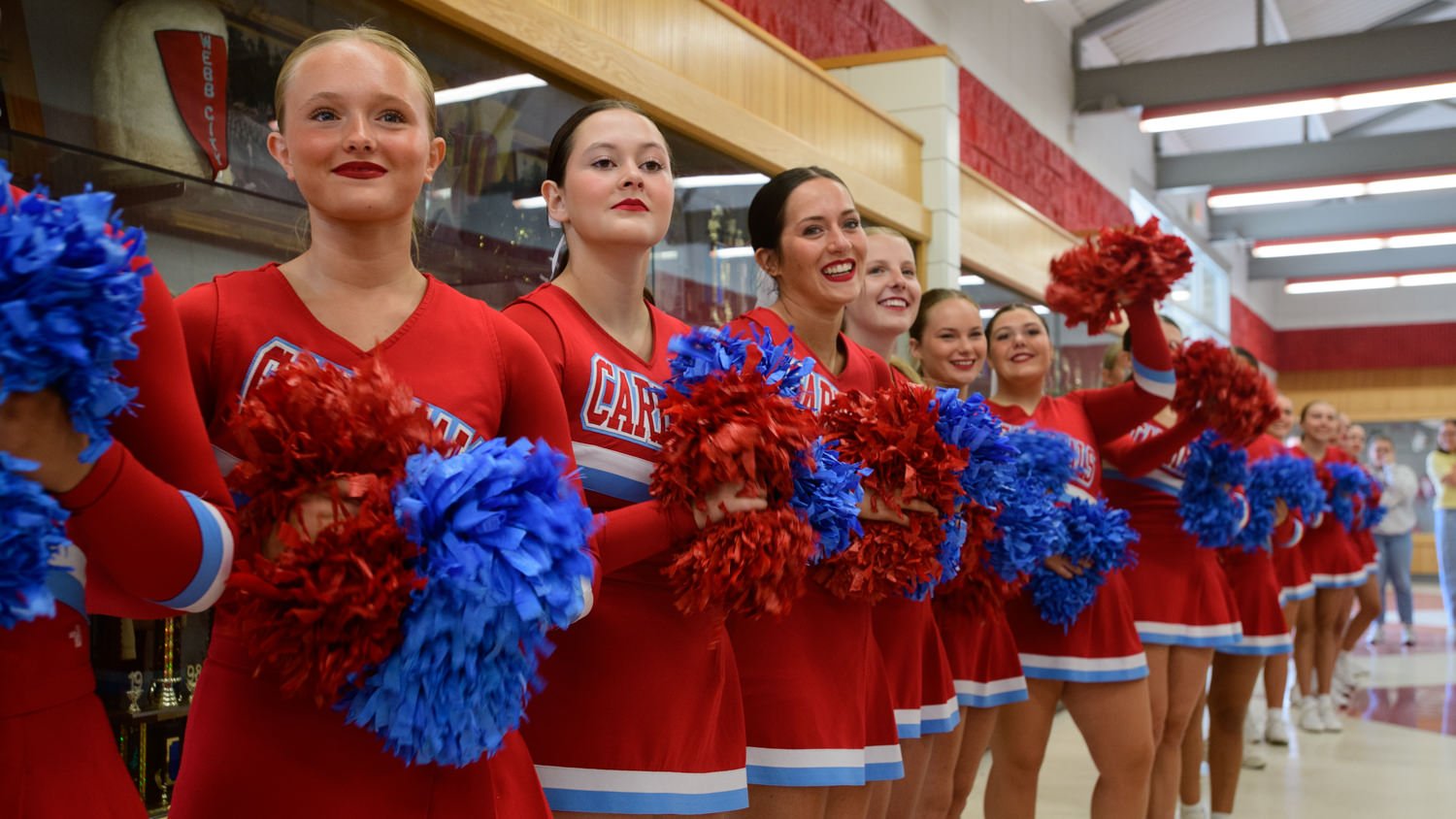 Line of cheerleaders in red uniforms holding red and blue pom-poms in a school hallway.