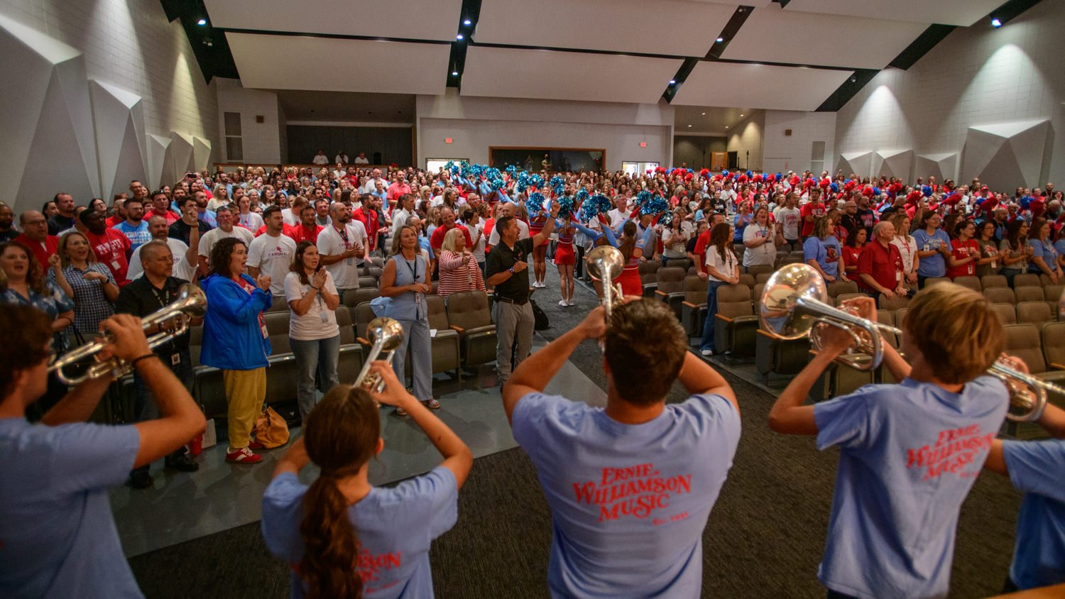 Band performs brass music to a cheering, large audience in a packed auditorium; crowd wears red, white, and blue shirts.