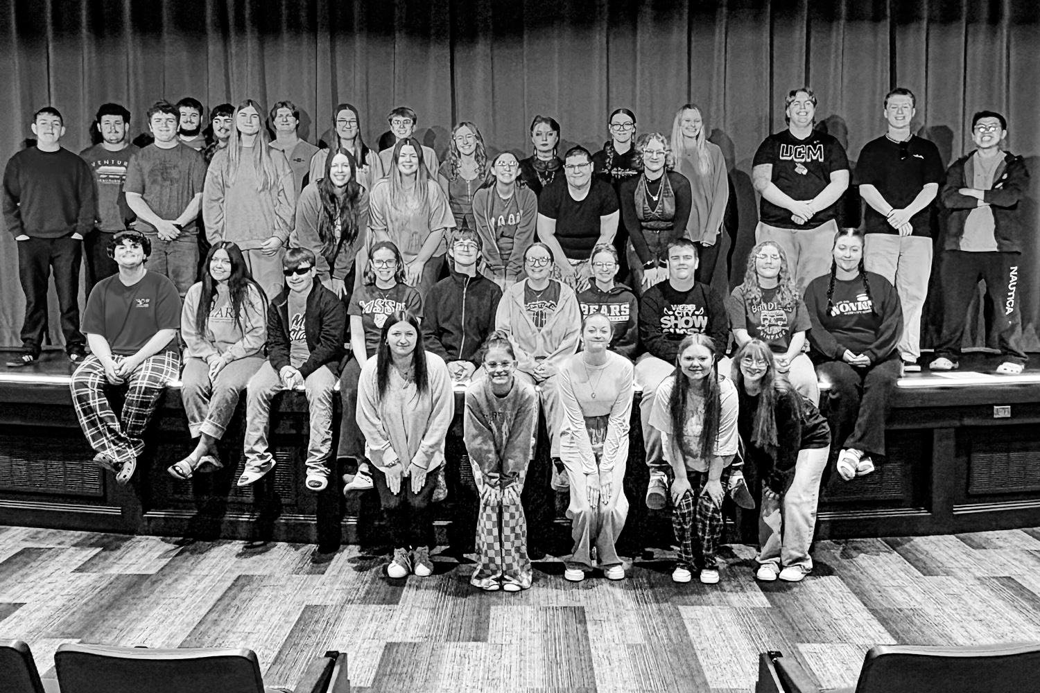 Large group of students posing on a stage in a theater, arranged in three rows and facing the camera.