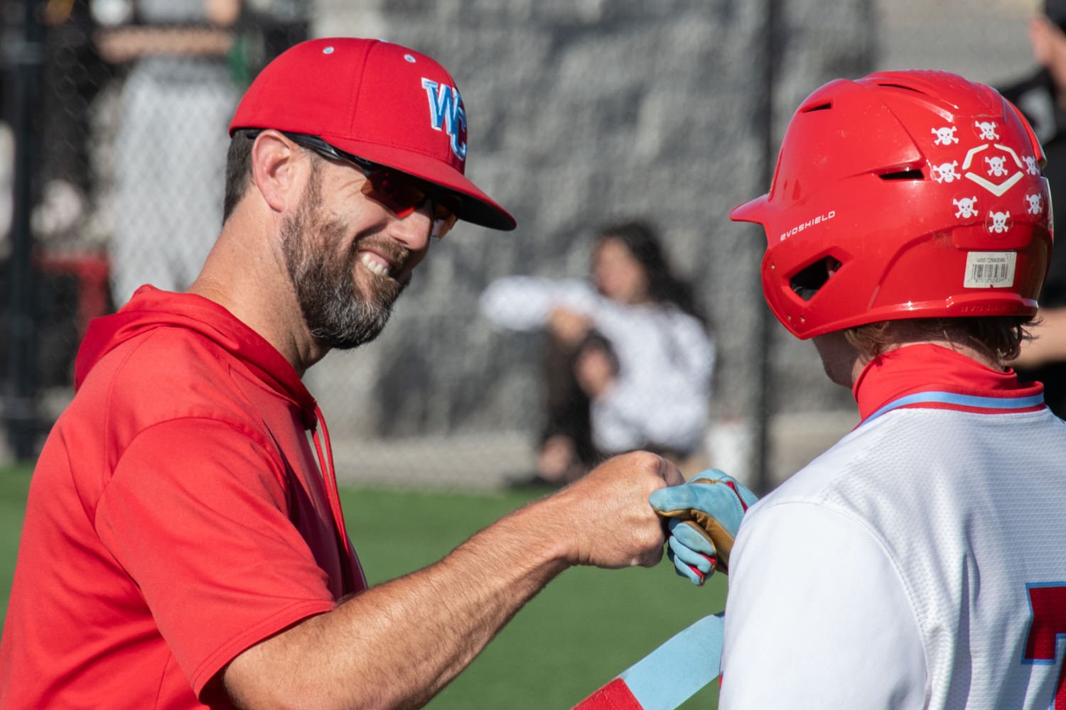 Coach in a red shirt and cap high-fiving a player in a red helmet on a baseball/softball field outdoors