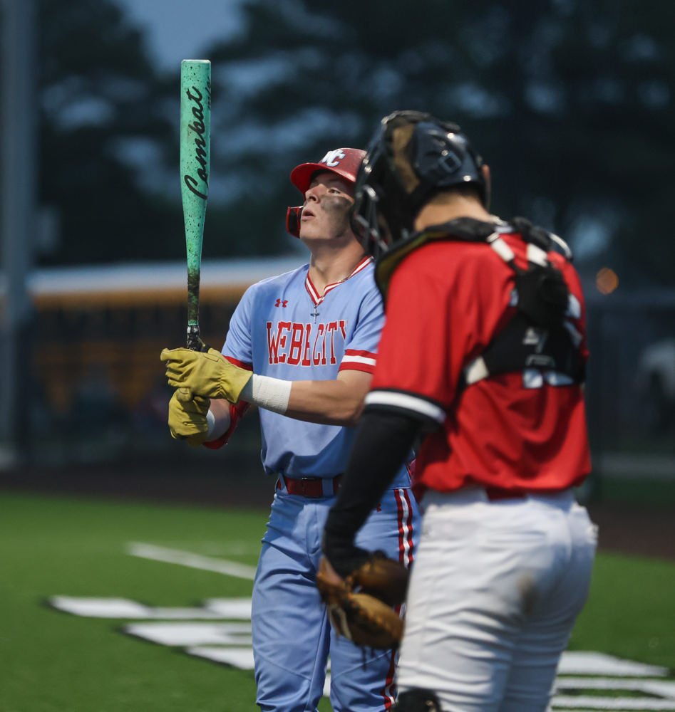 Baseball batter in a light blue uniform holds a green bat, looking up as he prepares to swing; catcher in red gear stands nearby.