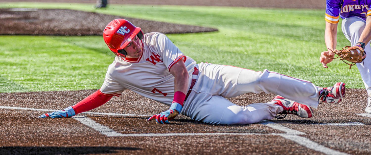 A baserunner slides headfirst into home plate in a white and red uniform with a red helmet, gloved hand reaching toward the bag.