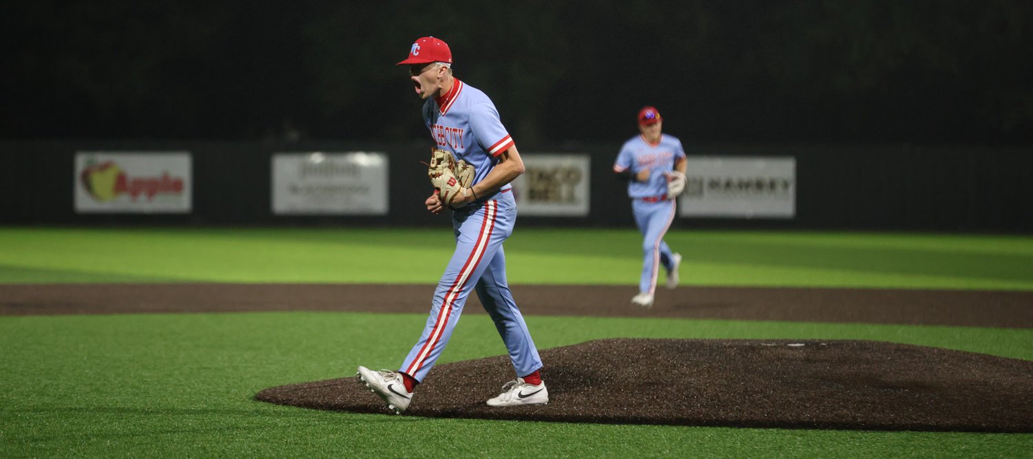 Baseball pitcher on the mound in a light-blue uniform with red cap, glove in hand during a night game; a fielder is visible in the background.