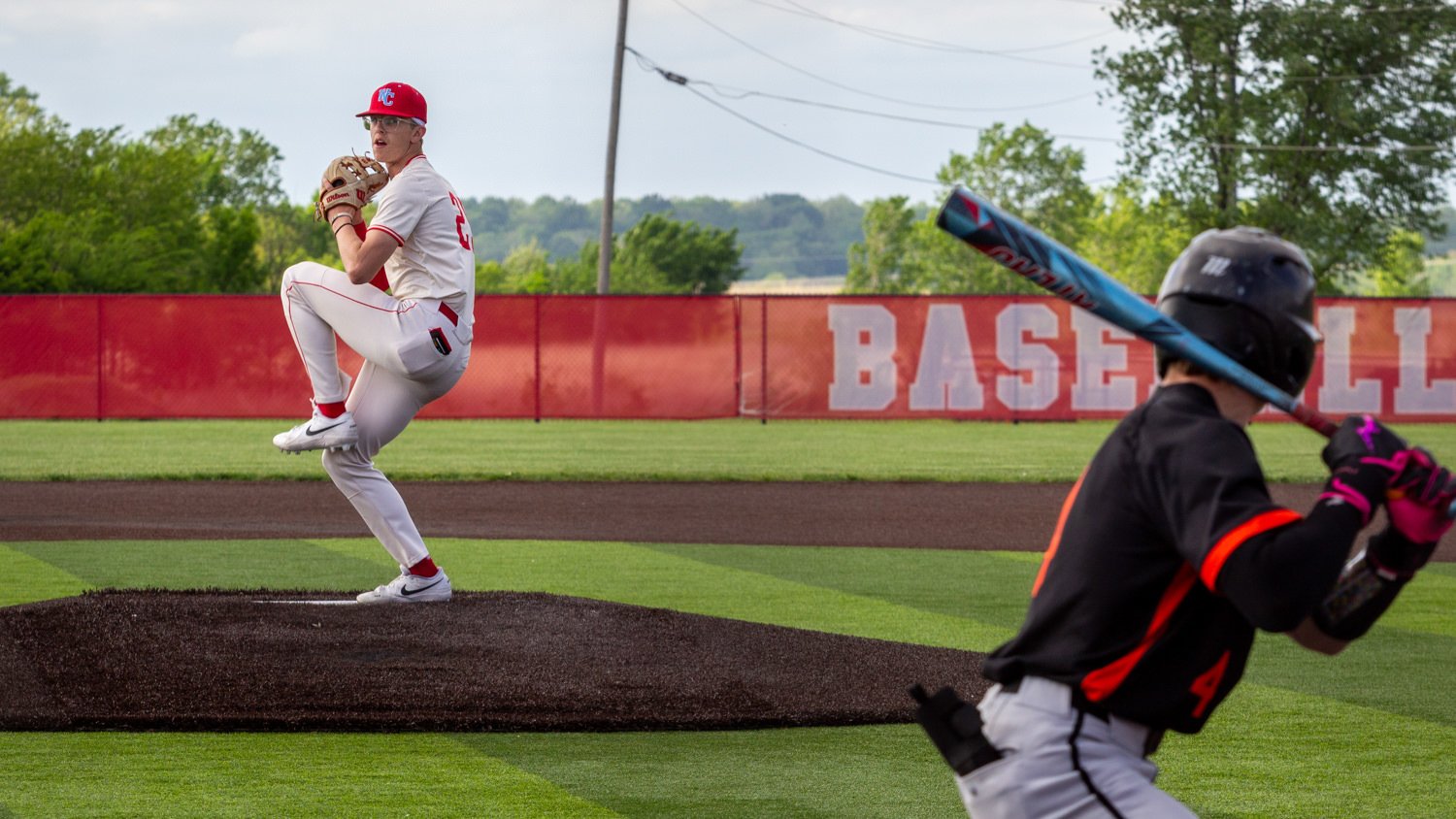 Baseball pitcher in white uniform on the mound delivering a pitch, with a batter in black and orange in the foreground.