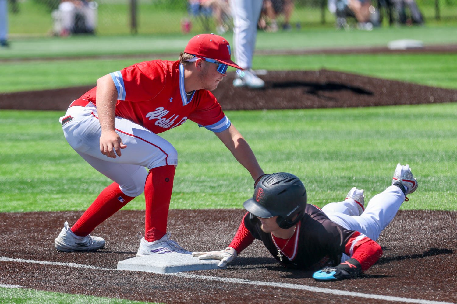 Baseball player in red reaches for a bag as a sliding opponent in black helmet and white pants attempts to reach it on a dirt basepath.