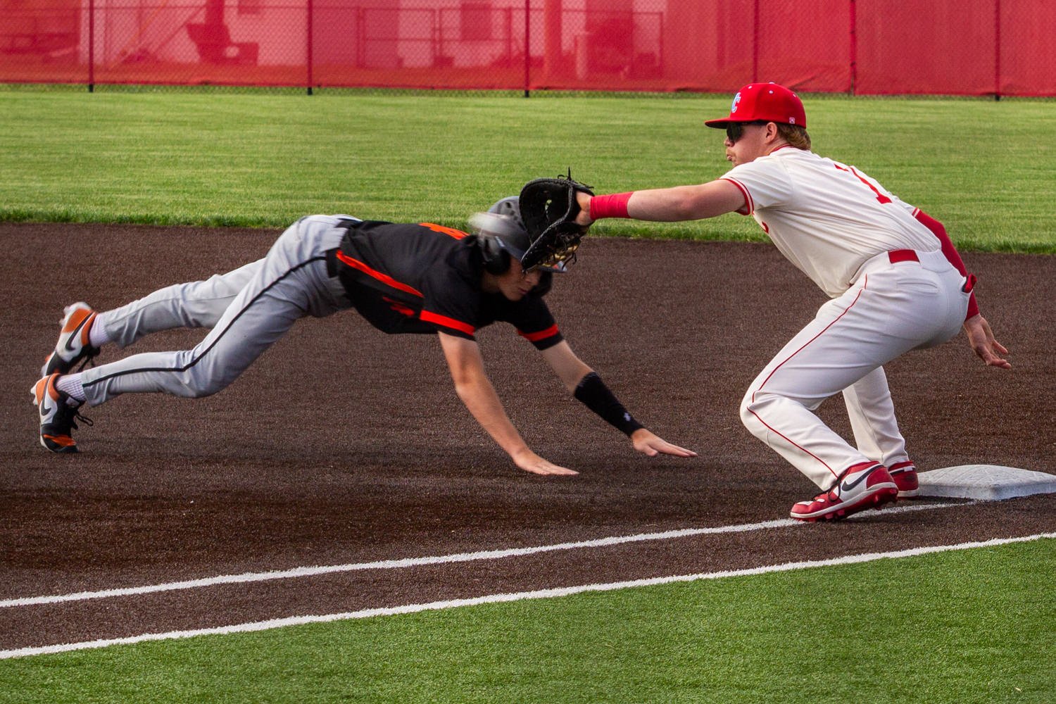 Baseball fielder in white reaches with his glove to tag a sliding baserunner near a base on a dirt infield edge.