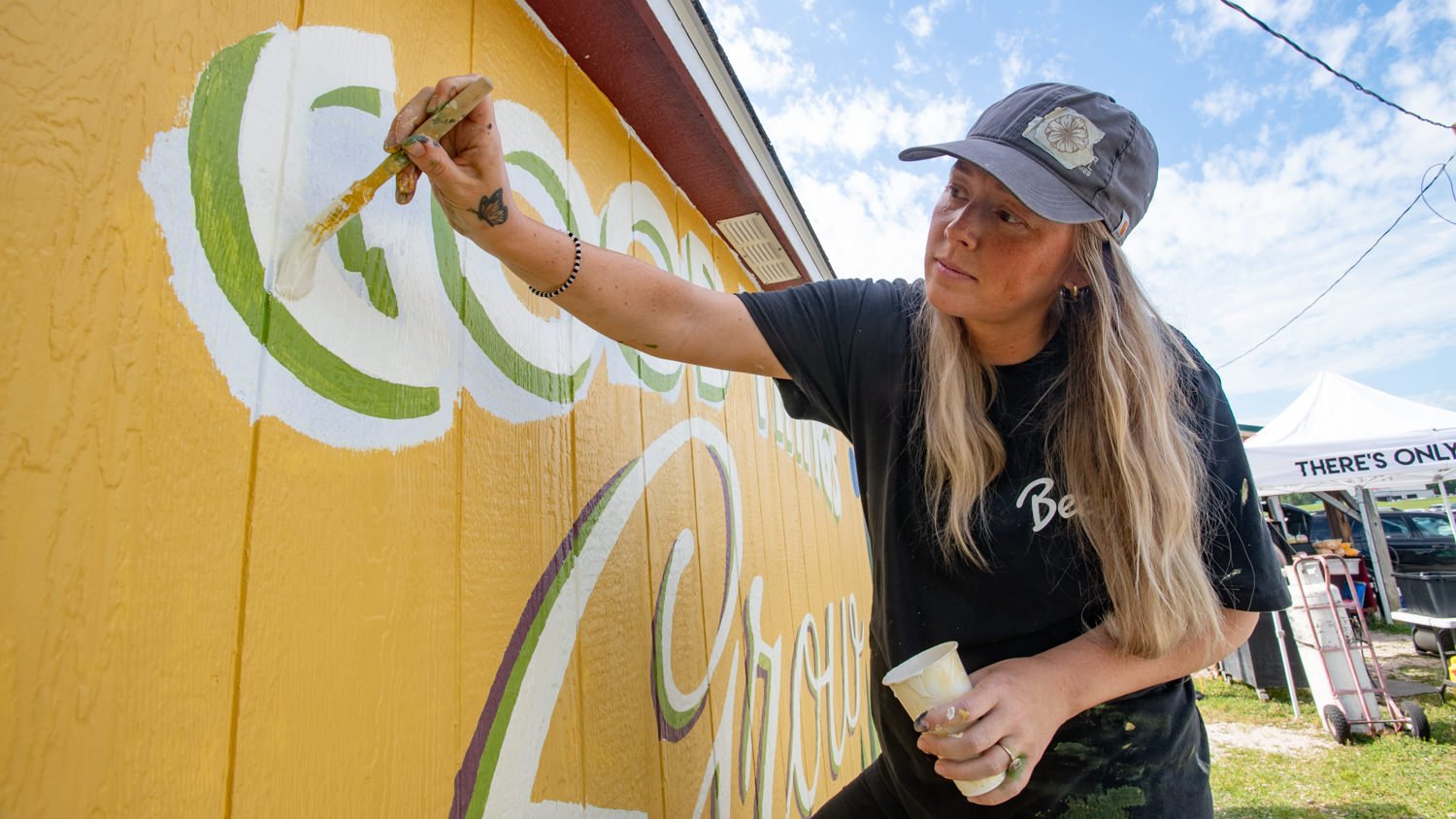 A woman wearing a cap and black shirt paints a colorful mural on a bright yellow wooden wall outdoors, holding a brush and cup.