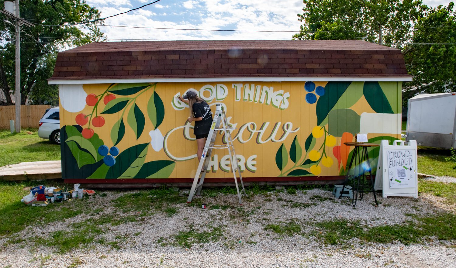 Person on a ladder painting a colorful mural on a small shed, with the words 'Good Things Grow Here' and fruit-and-leaf designs nearby.
