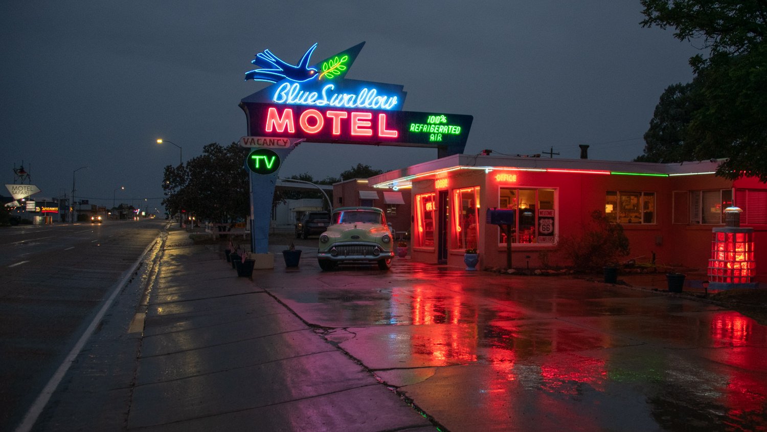 Neon-lit Blue Swallow Motel sign with a vintage car in front on a wet street at night, reflecting red and blue lights.
