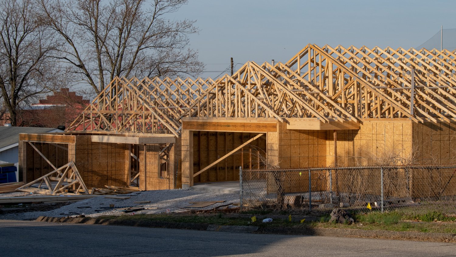 House under construction with exposed wooden frame and roof trusses, fenced yard and debris in the foreground