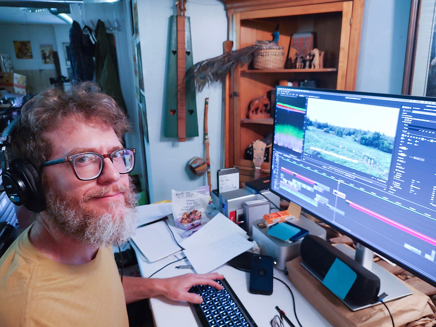 Man with glasses and a beard wearing over-ear headphones sits at a desk, editing video on a large monitor displaying a timeline and footage in a home office.