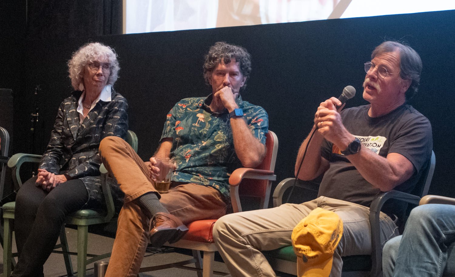 Three people sit on stage in a panel discussion; the man on the right speaks into a microphone while others listen.