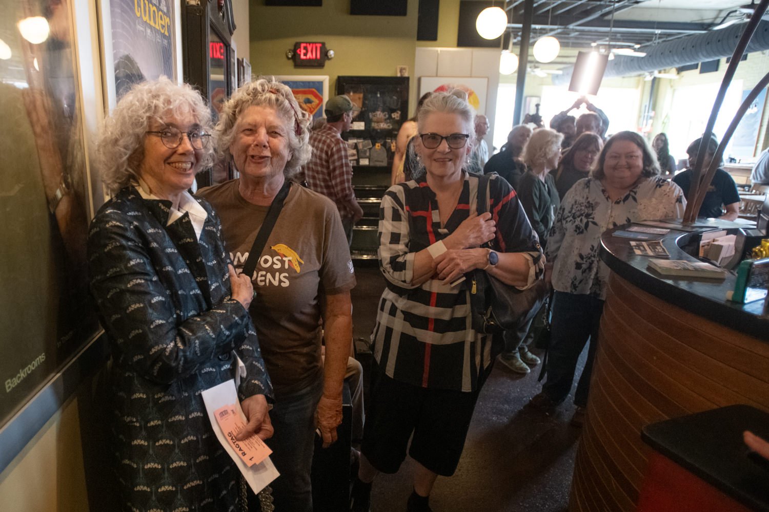 Three smiling women stand near a curved café counter inside a busy room, with other guests in the background.