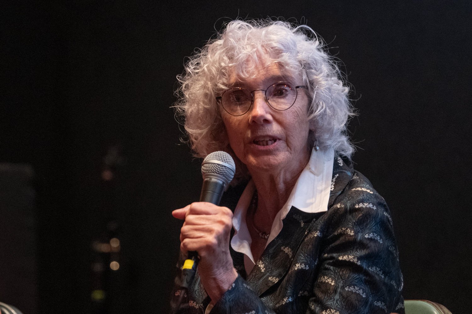 Elderly woman with curly white hair and glasses speaks into a handheld microphone on stage in a dark background.