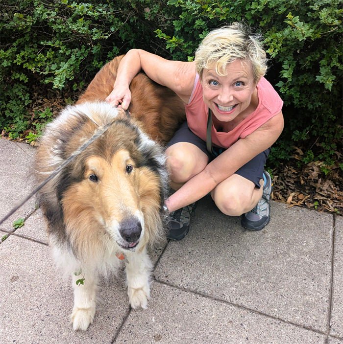 Smiling woman kneels on a sidewalk beside a large fluffy dog on a leash outside, both facing the camera.