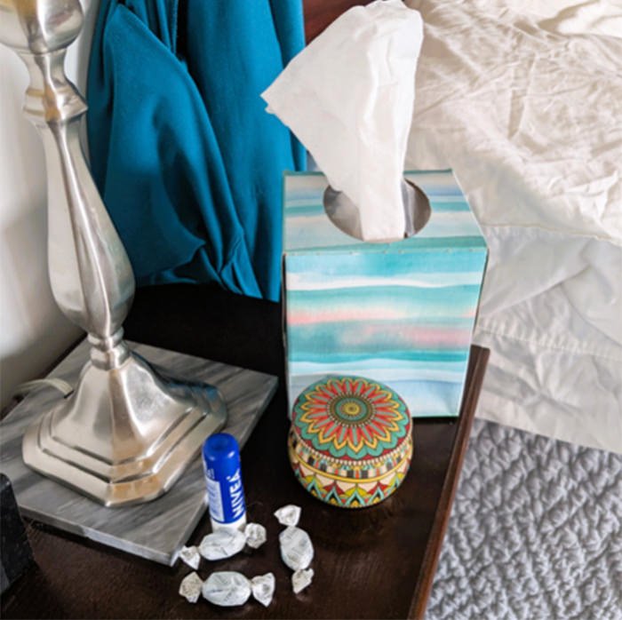 Nightstand with a tissue box, decorative round tin, lamp, blue scarf, and scattered candy wrappers near a blue glue stick.
