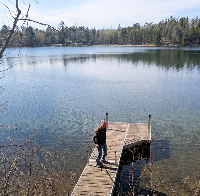 Older man in a brown jacket stands on a wooden dock extending into a calm lake, with forested shoreline in the background.