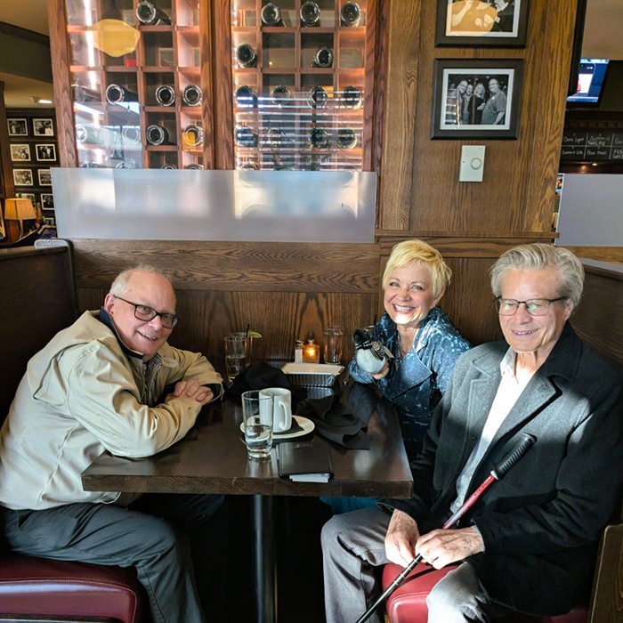 Three older adults sit at a restaurant table and smile at the camera; the woman in the middle holds a small black dog.