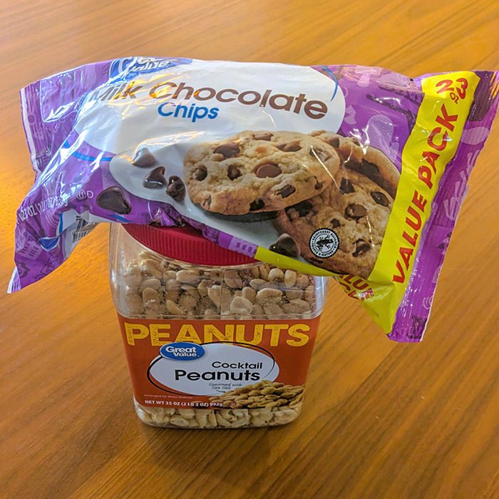 Jar of cocktail peanuts with a purple bag of milk chocolate chips resting on top, on a wooden table.