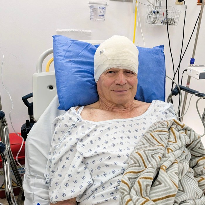 Older man in a hospital bed with a white head wrap, smiling, covered by a patterned blanket and blue pillows.