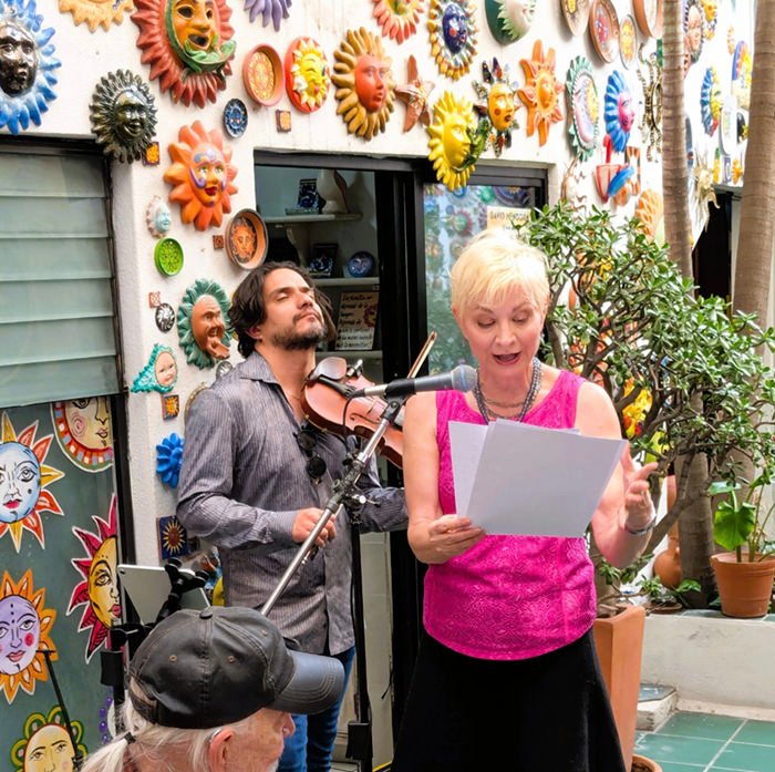 Woman in pink top sings from sheet music while a violinist accompanies outside a colorful sun-decorated wall.