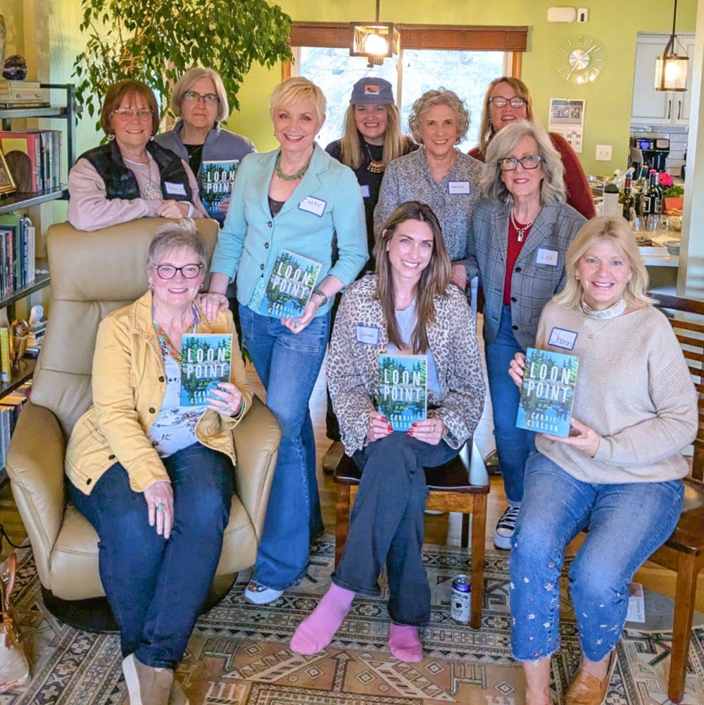 Nine women pose in a bright living room, each holding a copy of the book Loon Point and smiling.