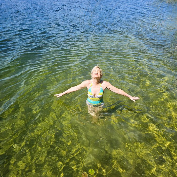Woman in a striped swimsuit stands chest-deep in clear green water with arms outstretched and eyes closed, enjoying a lakeside swim.
