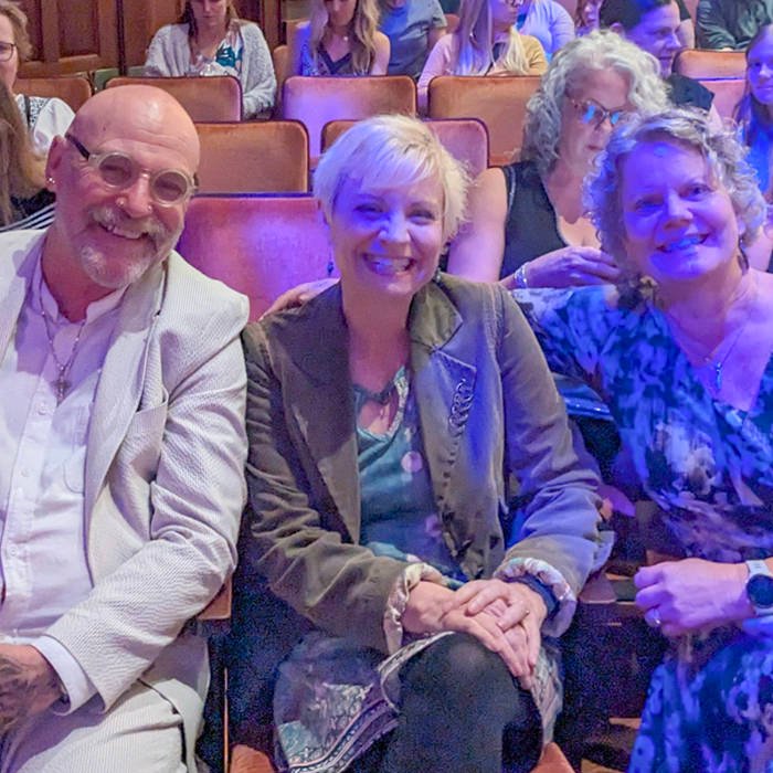 Three smiling theatergoers sit in row seats for a group photo, with others in the audience behind them.