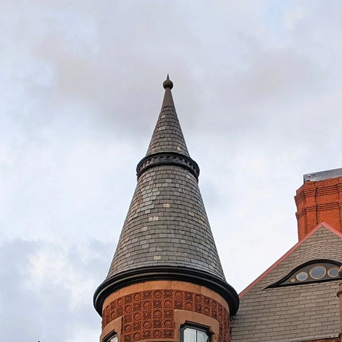 Conical slate-tiled turret on a red brick building against a cloudy sky.