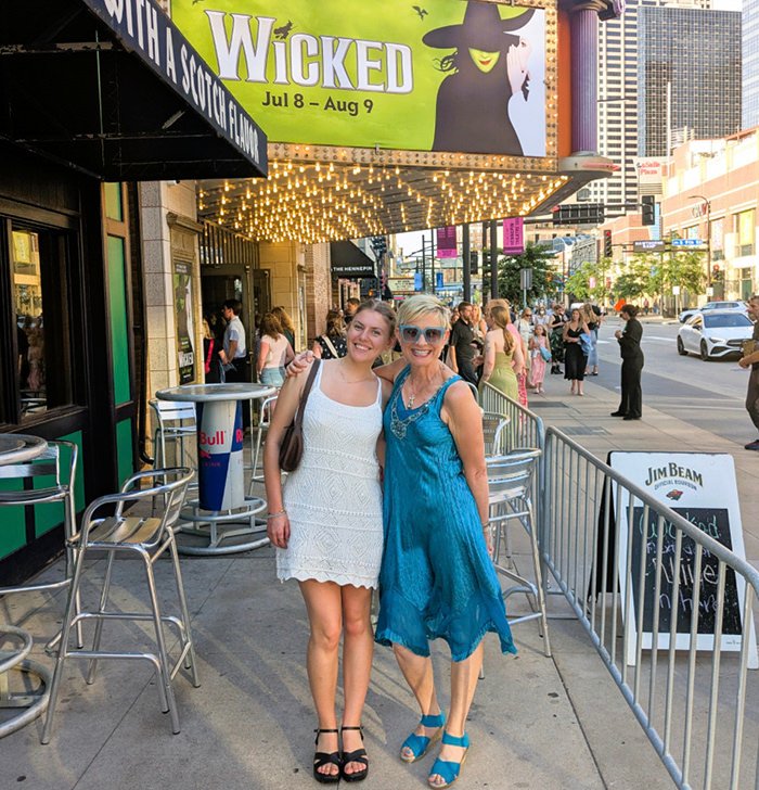 Two women pose together outside a theater marquee for Wicked, smiling at the camera.
