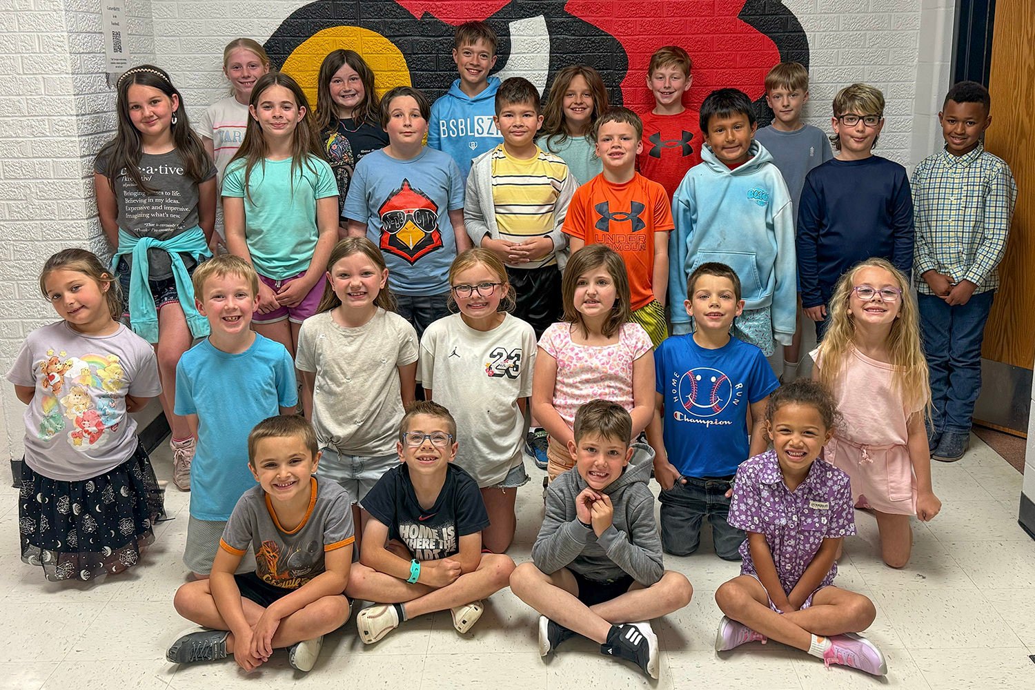 Group of about 24 elementary students posing for a class photo in a hallway with a colorful mural behind them.