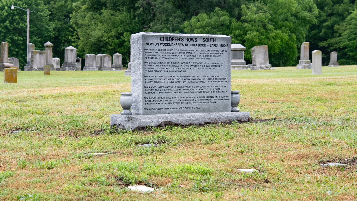 Granite memorial plaque in a grassy cemetery, titled 'Children's Rows – South,' with many headstones in the background.