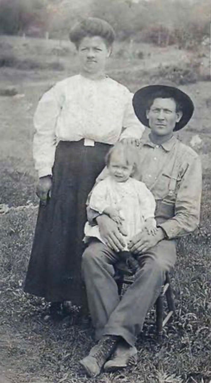 Man seated in a field holding a baby on his lap while a woman stands beside them outdoors (black-and-white family photo).