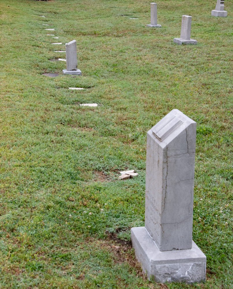 Grassy cemetery with a row of small upright white headstones, a larger rectangular tombstone leaning in the foreground.