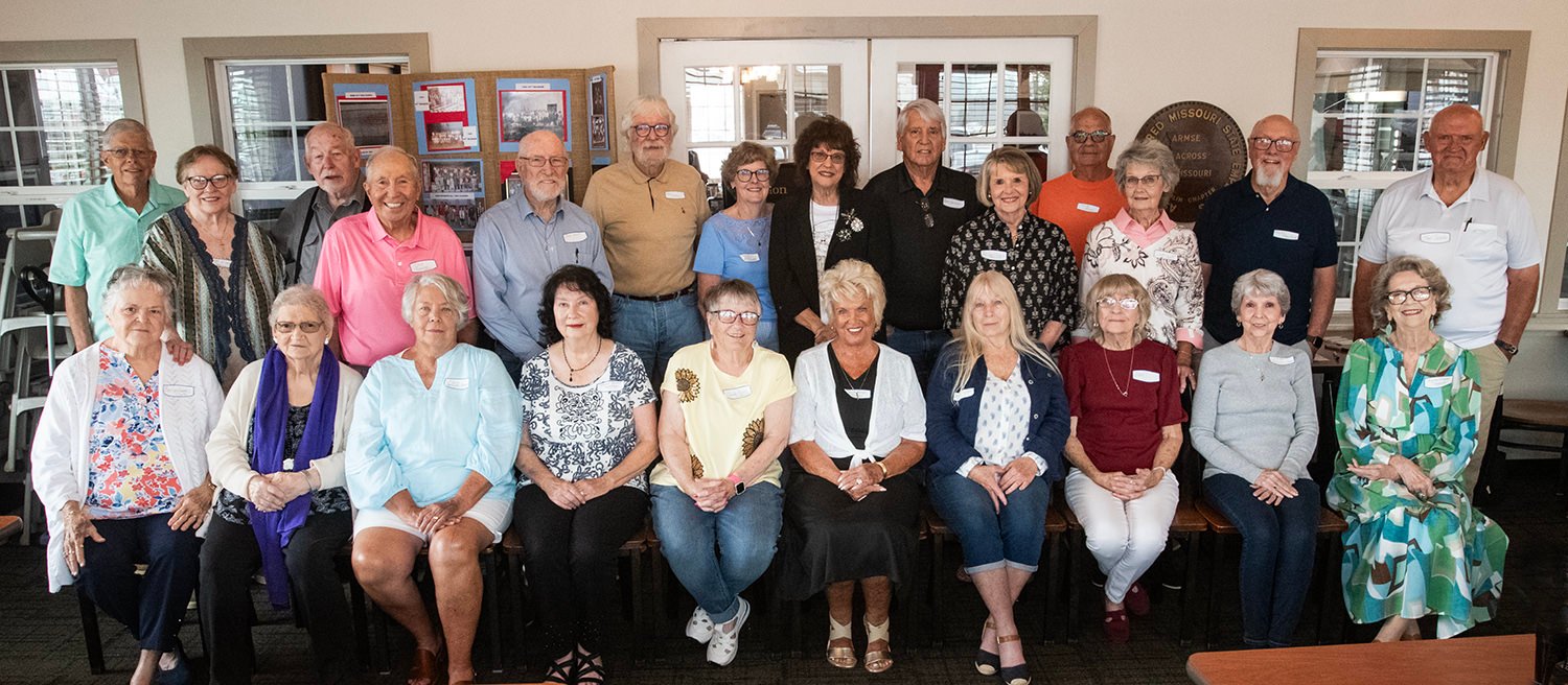 Group of adults, mostly seniors, posing for a group portrait in a community center with chairs and display boards in the background.